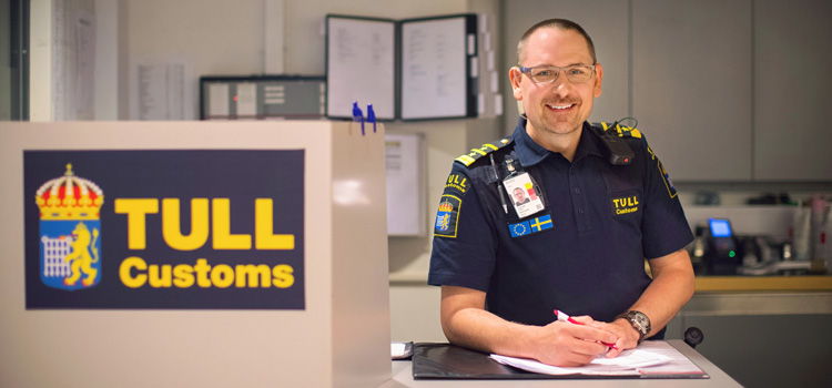Tullinspektör i uniform ler och står vid ett bord och skriver på en dokument.
